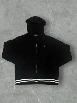 En svart zip hoodie från Ralph Lauren - En svart Ralph Lauren zip hoodie med vita streck där nere! Priset är självklart inte hugget i sten och är öppen för frågor om tröjan!
