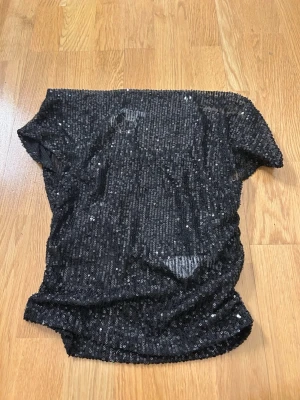 Svart paljettopp med kort ärm - Säljer en svart topp täckt med glittriga paljetter. Modellen har korta ärmar och är lätt transparent, vilket ger en cool och trendig look. Perfekt för dig som vill sticka ut och glänsa lite extra. Den kan ”fälla” lite. Men är super snygg 