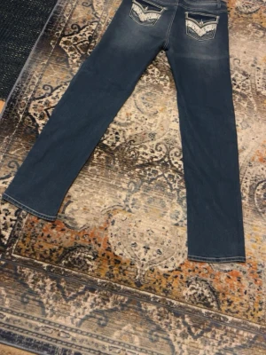 Blå jeans från Faded Glory med spets - Säljer ett par blå jeans från Faded Glory med raka ben och snygga detaljer på bakfickorna. Fickorna har vit spets och dekorativa knappar med glittrig sten. Jeansen har kontrastsömmar och klassisk femficksdesign. Perfekta för dig som gillar unika detaljer!