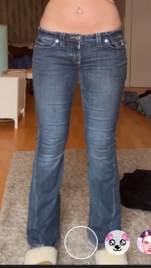 True Religion Joey Low Rise Flare Jeans - Säljer ett par blå True Religion Joey jeans med låg midja och flare-ben. Klassiska kontrastsömmar, stora fickor med lock och logga bak. Jeansen är i denim med snygg tvätt och detaljerade sömmar. Perfekt för dig som gillar en retro och avslappnad look. Fläck som troligen försvinner i tvätten, bild 1, syns knappt. Även trådar som lossnat där man knäpper byxorna, men inget som gör att man inte kan stänga byxorna osv. Säljer billigare pga defekterna orginalpris 1299