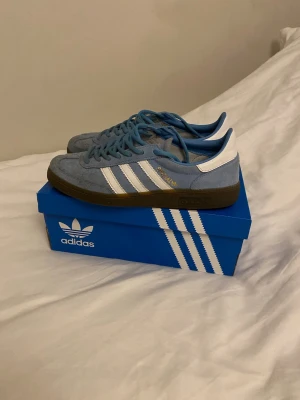 Adidas Handball Spezial ljusblå sneakers - Säljer ett par Adidas Handball Spezial sneakers i ljusblå mocka med vita detaljer och klassiska tre ränder på sidan. Skorna har bruna gummisulor, blå snörning och gulddetaljer på sidan och tungan. Perfekta för dig som gillar retrostil och streetwear.