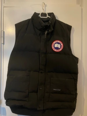 Svart dunväst från Canada Goose - Säljer en svart dunväst från Canada Goose med klassisk logga på bröstet. Västen har hög krage, två stora fickor framtill och stängs med både dragkedja och knappar. Perfekt för lager-på-lager och riktigt varm under kyliga dagar.