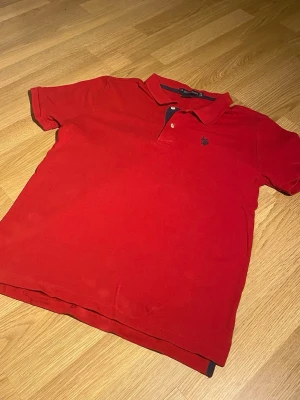 Röd pikétröja från U.S. Polo Assn - Klassisk röd pikétröja från U.S. Polo Assn med krage, två knappar och mörkblå broderad logga på bröstet. Tillverkad i mjuk bomull och har korta ärmar. Perfekt till jeans eller shorts för en avslappnad look.