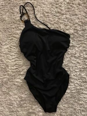 Svart baddräkt med ringdetalj - Säljer en svart baddräkt med asymmetrisk axel och cool ringdetalj i plast på ena axelbandet. I storlek XS. Baddräkten har tunna axelband och är tillverkad i ett stretchigt syntetmaterial som sitter snyggt på kroppen. Perfekt för stranden eller poolen! Gåva vid köp!🎁