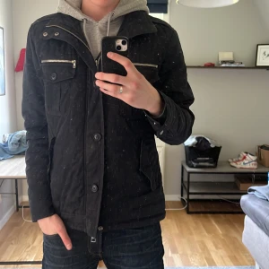 Fieldjacket från Riley med dragkedjor - Mörkblå field jacket från Riley med snygga silverfärgade dragkedjor på bröstfickorna och knappar på fickor och ärmslut. Jackan har hög krage, flera fickor och en stilren look. Perfekt för dig som vill ha en clean och funktionell jacka med lite edge. Finns även en luva man kan dra ut från kragen