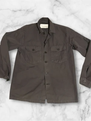Grå grön overshirt från A days march - Säljer en oliv overshirt med klassisk krage och knäppning framtill. Jackan har två stora bröstfickor med lock och knappar, samt långa ärmar. Perfekt att slänga över en hoodie eller t-shirt för en chill look.