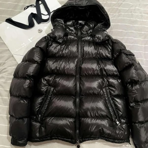 Moncler maya - Moncler Maya puffjacka i storlek M/2 och svart färg. Tillverkad i blank nylon med gåsdunfyllning som håller dig varm och bekväm. Jackan har en tvåvägsdragkedja, avtagbar huva, fickor med dragkedjor och Moncler logga på armen. Mycket fint skick och perfekt för nästan alla tillfällen.
