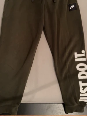 Olivgröna mjukisbyxor från Nike  - Säljer ett par olivgröna mjukisbyxor från Nike i storlek S. Byxorna har resår och snörning i midjan, samt trycket 'JUST DO IT.' i vitt längs ena benet. Tillverkade i mjuk bomullsmix för maximal komfort. Perfekta för chill eller träning.