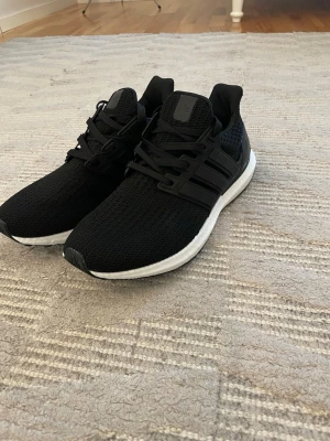 Svarta Adidas Ultraboost sneakers - Säljer ett par svarta Adidas Ultraboost sneakers med vit sula och svart ovandel i mesh. Skorna har snörning, Torsion System och Continental-gummisula för extra grepp. Perfekta för dig som vill ha både stil och komfort till vardags eller träning.helt nya