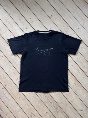 Vintage Y2K Nike svart t-shirt med tryck - Nike t-shirt i svart med ett tryck av Nike-loggan på framsidan. Lappen borta men estimerad storlek är L.  I fint skick förutom att sömmen gått upp något nere i hemmen, se bild. Går att snabbt sy för hand eller symaskin. Annars inget som syns eller påverkar tröjans funktion.