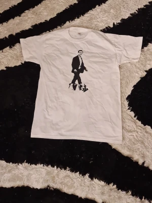 Vit t-shirt med svart tryck - Vit t-shirt från Fruit of the Loom med ett svart tryck av en person i kostym på framsidan. Klassisk rund halsringning och korta ärmar. Perfekt för dig som gillar enkel stil med en twist.