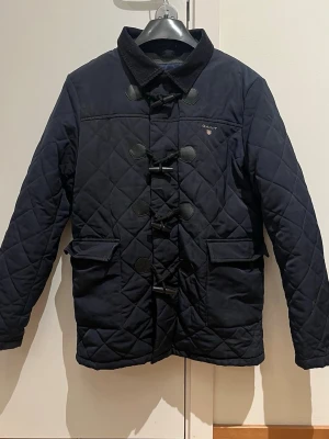 Mörkblå quiltad jacka från Gant - Säljer en mörkblå quiltad jacka från Gant med snygga träknappar och snörning framtill. Jackan har två stora fickor, mjukt fleecefoder och krage i manchester. Perfekt för kyliga dagar och riktigt skön att bära. Storlek 158/164 (xs)