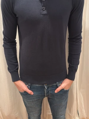 Mörkblå långärmad pikétröja - Säljer en stilren mörkblå långärmad pikétröja med knappslå framtill. Tröjan har en slim passform och är tillverkad i mjuk bomull som känns skön mot huden. Perfekt till jeans för en clean och enkel look. Märket är indicode och storlek S