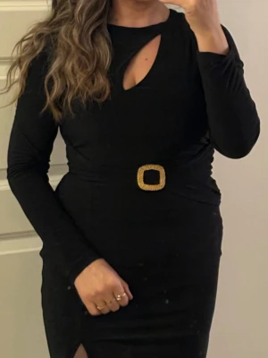 Svart långärmad klänning med guldspänne - Snygg svart långärmad klänning med ett dekorativt guldspänne i midjan och en liten cut-out vid halsen. Klänningen har en slits framtill och är figurnära i modellen. Perfekt för dig som vill ha en elegant och stilren look.