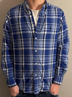 Blårutig skjorta från Ralph Lauren - Säljer en klassisk blårutig skjorta från Ralph Lauren med vit knappstängning och broderad logga på bröstet. Skjortan har lång ärm, button-down krage och är tillverkad i mjuk bomull. Perfekt för dig som gillar stilrena och tidlösa plagg.