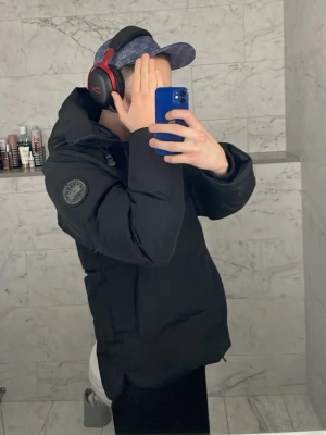 Svart pufferjacka från Canada Goose - Säljer en svart pufferjacka från Canada Goose med huva och diskret logga på ärmen. Jackan är vadderad och har en clean look med dragkedja och knappar framtill. Perfekt för kalla dagar och riktigt skön att bära. Snygg och enkel att matcha med allt.