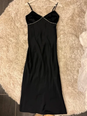 Svart långklänning med strassdetaljer - Supersnygg svart lång satinklänning med tunna axelband och glittrande strassdetaljer längs bysten. Se bild för tappade stenar, inget som syns tydligt. 