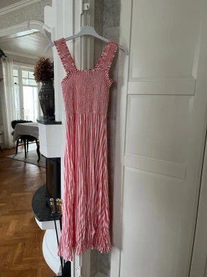 Randig långklänning från Pieces  - Superfin långklänning från Pieces i vitt och peach röd med breda axelband och smock upptill. Klänningen har ett randigt mönster och är gjord i ett lätt bomullstyg, perfekt för varma sommardagar. Den är ej struken nertill så därför är den lite skrynklig!