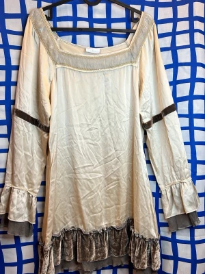 Ljus volangklänning från Valerie - Unik ljus klänning från Valerie med volangdetaljer i sammet och mesh vid ärmslut och nederkant. Långärmad modell med dekorativa band i sammet på ärmarna och guldfärgade detaljer vid halsen. Perfekt för dig som gillar romantisk och bohemisk stil.