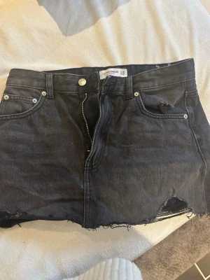 Svart jeanskjol från Pull&Bear - Säljer en svart jeanskjol från Pull&Bear med rå kant och slitningar. Kjolen har klassiska fem fickor, bälteshällor och dragkedja framtill. Perfekt för dig som gillar en edgy och avslappnad stil.