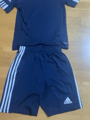 Adidas marinblått träningsset - Säljer ett marinblått träningsset från Adidas med klassiska vita ränder på ärmar och ben. T-shirten har Adidas-logga på ärmen och byxorna har loggan på benet. Materialet är lätt och andas, perfekt för sport eller träning.