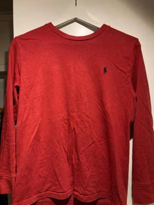 Röd långärmad tröja Polo Ralph Lauren - Röd långärmad tröja från Polo Ralph Lauren i mjuk bomull. Klassisk rund halsringning och ikoniska mörkblå polospelaren broderad på bröstet. Perfekt till jeans eller chinos för en clean och stilren look.