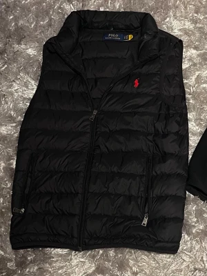 Svart dunväst från Polo Ralph Lauren - Säljer en svart dunväst från Polo Ralph Lauren med klassisk röd logga på bröstet. Västen har hög krage, två fickor med dragkedja och quiltad design. Perfekt att slänga över en skjorta för en clean och stilren look.