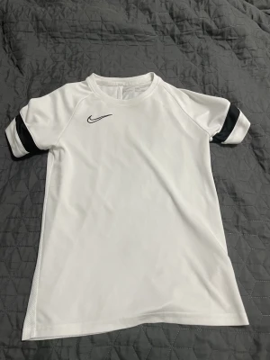 Vit Nike Dri-Fit t-shirt - Säljer en clean vit Nike Dri-Fit t-shirt med svart broderad swoosh på bröstet och svarta detaljer på ärmarna. Tillverkad i lätt och ventilerande polyester som passar perfekt till träning eller sport. Klassisk passform och rund hals.