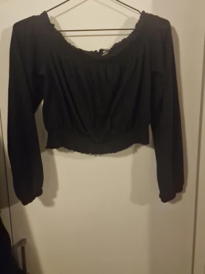 Svart croppad blus med lång ärm - Säljer en svart croppad blus med långa ärmar och resår vid midjan och ärmsluten. Toppen har en söt offshoulder-ringning och lätt puffiga ärmar som ger en trendig look. Perfekt att styla med höga jeans eller kjol.