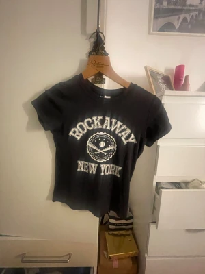 Svart Rockaway t-shirt - Säljer en svart t-shirt från Divided med trycket 'Rockaway New York' och ett coolt basebollmotiv framtill. T-shirten har korta ärmar och en klassisk rund hals. 