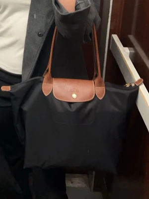 Svart shoppingväska från Longchamp - Klassisk svart shoppingväska från Longchamp med bruna skinnhandtag och lock. Väskan har guldiga detaljer och dragkedja upptill. Perfekt storlek för att få plats med allt du behöver och enkel att bära över axeln.