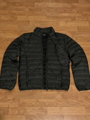 Mörkgrön pufferjacka med dragkedja - Säljer en mörkgrön pufferjacka med horisontella sömmar och hög krage. Jackan har dragkedja framtill och två sidofickor med dragkedja. Lätt och smidig modell som är enkel att packa ner i medföljande påse.