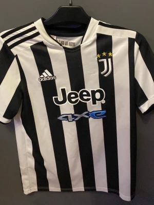 Juventus adidas matchtröja 10-12 år. - Juventus matchtröja från adidas i svartvitt randigt mönster med klubbmärke, tre stjärnor och Jeep-logga på bröstet. Tillverkad i Primegreen-material och har klassiska adidas-ränder på axlarna. Perfekt för fotbollsfans!