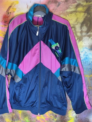 Lotto vintage track jacket 90-tal retro jacka L - Snygg och äkta vintage track jacket från Lotto i klassisk 90-talsdesign. Jackan har en mörkblå bas med rosa, turkosa och neongröna detaljer som verkligen sticker ut. Broderad Lotto-logga på bröstet och originaldragkedja med Lotto-zip. Tillverkad i 100 % polyester och producerad i Malaysia, vilket är typiskt för äldre Lotto-plagg. Passformen är boxig och tidstypisk, storlek L enligt etikett och funkar även oversized beroende på hur man vill bära den.