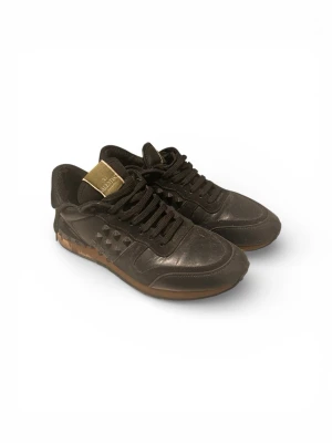Valentino Rockrunner svarta sneakers - Svarta Valentino Rockrunner sneakers i skinn med coola nitar på sidorna och klassisk snörning. Skorna har en brun sula och gulddetalj på plösen med Valentino-logga. Perfekta för dig som vill ha en edgy och lyxig look.