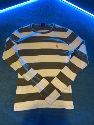 Randig långärmad tröja Ralph Lauren - Säljer en klassisk långärmad tröja från Ralph Lauren Sport i vitt och mörkgrönt med breda ränder. Tröjan har rund halsringning och den ikoniska orangea Polo-loggan broderad på bröstet. Tillverkad i 100% bomull för en skön och avslappnad look. Fläckar finns men de går att tvätta 