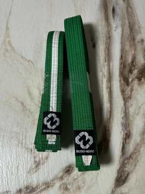 Grönt bälte Budo-Nord 240/300cm - Två gröna bälten från Budo-Nord, ett med vit rand och ett helgrönt. Tillverkade i slitstarkt tyg, 55% bomull och 45% polyester på det längre bältet. Perfekt för kampsportsträning och passar dig som vill levla upp din look på mattan.