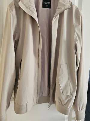 Beige vindjacka från Peak Performance - Snygg beige vindjacka från Peak Performance i storlek L. Jackan har dragkedja, höga fickor med lock och ribbade muddar vid ärmslut och nederkant. Perfekt för vår och höst, enkel och stilren design som passar till det mesta.