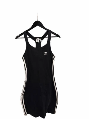 Svart sportig klänning från Adidas - Snygg svart kortklänning från Adidas med vita ränder längs sidorna och klassisk logga på bröstet. Klänningen har breda axelband och ärmlös design