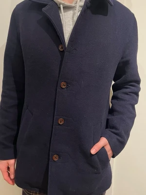 Mörkblå rock från Percival - Säljer en mörkblå rock från Percival med klassisk krage och snygga bruna knappar. Yttertyget är i en mix av ull och polyester, fodrad med randigt bomullstyg. Perfekt för dig som vill ha en stilren och tidlös jacka till höst och vinter.