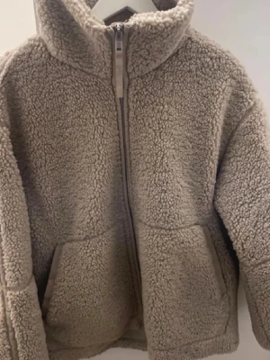 Beige teddyjacka med dragkedja - Mysig beige teddyjacka med hög krage och hel dragkedja framtill. Jackan har två fickor och är tillverkad i fluffigt fleece-material som håller dig varm under kyliga dagar. Perfekt för dig som gillar en avslappnad och trendig stil.