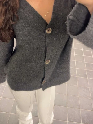 Mörkgrå stickad kofta med knappar - Mysig mörkgrå stickad kofta med stora träknappar framtill. Koftan har en avslappnad passform och långa ärmar, perfekt att slänga på sig under kyliga dagar. Enkel och stilren design som funkar till det mesta.