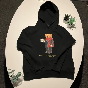 Svart hoodie med björnmotiv från Ralph Lauren - Säljer en svart hoodie från Ralph Lauren med ett charmigt björnmotiv på framsidan. Björnen har en röd halsduk och håller en kopp med rykande dryck. Perfekt för en avslappnad stil med en touch av elegans. 🐻☕lite nopprig men annars väldigt fin (OPS! Ganska liten i storleken!😃)
