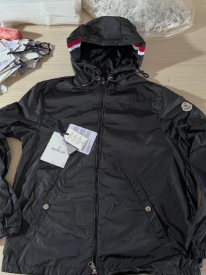 Svart vindjacka från Moncler - Säljer en svart vindjacka från Moncler med huva och dragkedja framtill. Jackan har Moncler-logga på ärmen och snygga detaljer med röd och vit rand på insidan av huvan. Tillverkad i lätt och glansig polyester, perfekt för dig som vill ha en clean och sportig look.