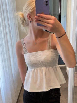Vit linnetopp med spetsband - Säljer en söt vit linnetopp med breda spetsband och smockad överdel. Toppen har en volang nedtill som ger en luftig och somrig känsla. Perfekt att matcha med jeans eller kjol för en chill look. Aldrig använd!