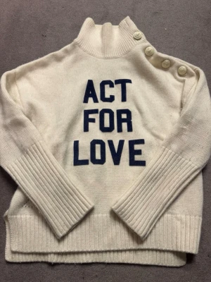 Vit stickad tröja Zadig & Voltaire - Säljer en vit stickad tröja från Zadig & Voltaire med texten 'ACT FOR LOVE' i mörkblått framtill. Tröjan har hög krage, ribbade muddar och fyra dekorativa knappar på axeln för en extra snygg detalj. Perfekt för dig som gillar statement-plagg.