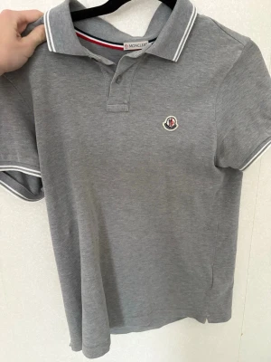 Grå Moncler pikétröja - Säljer en stilren grå pikétröja från Moncler med klassisk krage och knappar framtill. Tröjan har vita detaljer på krage och ärmslut samt Moncler-logga broderad på bröstet. Perfekt till jeans eller shorts för en clean look.