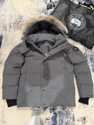 Grå dunjacka från Canada Goose - Säljer en grå dunjacka från Canada Goose med avtagbar huva och pälsdetalj. Jackan har svarta muddar, tryckknappar och dragkedja framtill samt klassisk patch på ärmen. Perfekt för kalla vinterdagar och riktigt snygg passform.