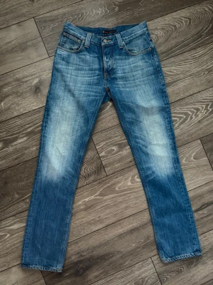 Nudie grim Tim - Säljer ett par klassiska blå jeans från Nudie Jeans med snygg slitning och kontrastsömmar. Modellen är grim Tim och har raka ben, fem fickor och ikonisk broderad detalj på bakfickorna. Tillverkade i slitstarkt bomullstyg och har normal passform.