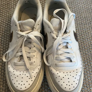 Nike Air Force 1 med leopard swoosh - Säljer ett par vita Nike Air Force 1 sneakers med leopardmönstrad swoosh och guldfärgade Nike-detaljer på hälen. Skorna har klassisk perforerad tå, snörning och är tillverkade i skinn. Perfekta för dig som vill ha en unik twist på dina sneakers.